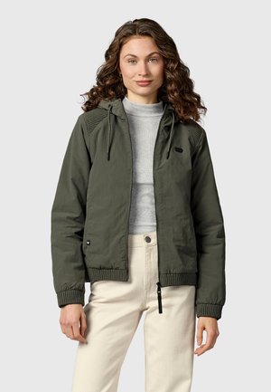 Kobieta o kręconych włosach, ubrana w oliwkowo-zieloną kurtkę z kapturem na suwak, na jasnoszarym golfie i beżowych spodniach, stojąca na jednolitym tle.