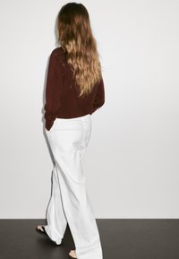 Burgundské pletené svetlo, bílé široké kalhoty, uvolněný střih. Dlouhé vlnité hnědé vlasy, viděné ze zadu, minimalistické pozadí.