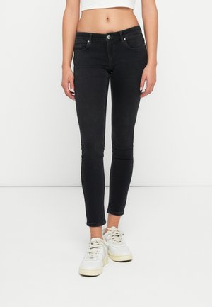 Femme portant un jean skinny noir taille haute et des baskets blanches, debout sur un fond blanc uni.