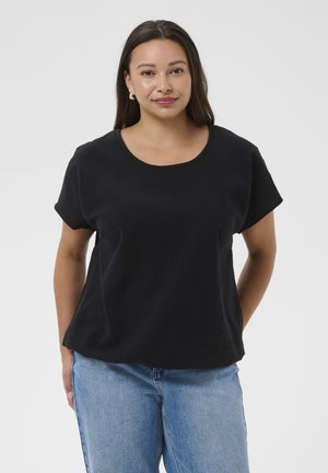 Frau mit langen dunklen Haaren, die Perlenohrringe, ein schwarzes kurzärmeliges Oberteil und hellblaue Jeans trägt, stehend vor einem schlichten hellen Hintergrund.