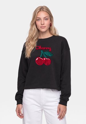 Lyshåret kvinde iført en sort sweater med røde kirsebær og teksten "Cherry", kombineret med hvide bukser mod en ensfarvet lys baggrund.