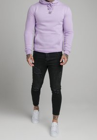 Sudadera de lavanda con bolsillo frontal y capucha con cordón, combinada con jeans skinny negros desgastados y zapatillas blancas.