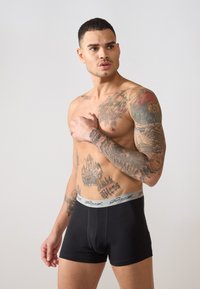 Fekete boxertati szürke rugalmas derékpánttal, amelyen aláírás logó található. Az anyag simának és testre simulónak tűnik, kiemelve a tetoválásokat.