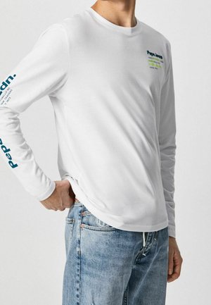 Homme portant une chemise blanche à manches longues avec un texte bleu sur la poitrine et la manche, associée à un jean en denim bleu clair, debout devant un fond uni.