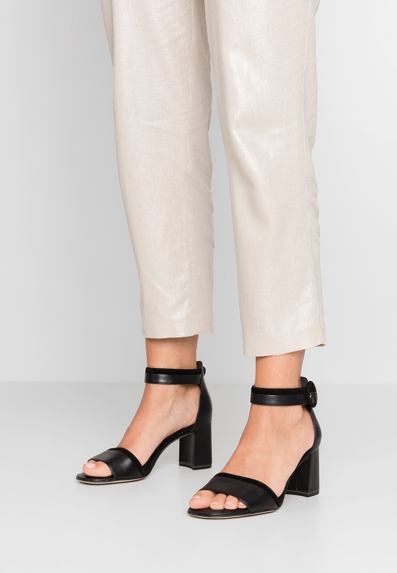 Sandales à talons carrés en cuir noir à bout ouvert avec brides à la cheville, portées avec un pantalon beige clair raccourci sur un sol gris.