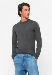 Pull gris à manches longues en maille, col rond, arborant un petit logo sur la poitrine, associé à un jean en denim bleu clair.