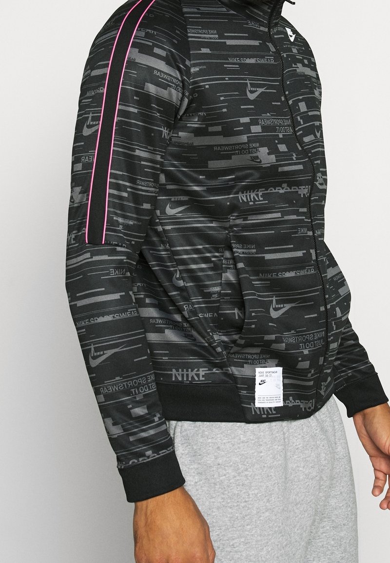 Svart zip-up jacka med Nike-logotyp, med en texturerad mönster, rosa ränder på axlarna och framfickor. Tillverkad av ett slätt material.