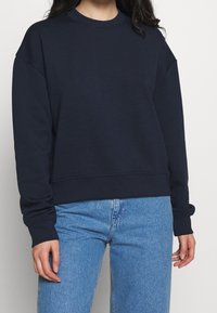 Felpa blu navy realizzata in tessuto morbido, con scollatura a girocollo, spalle scese, polsini a coste e orlo corto. Abbinata a jeans in denim chiaro.