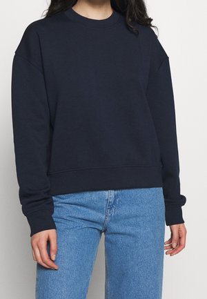 Felpa blu navy realizzata in tessuto morbido, con scollatura a girocollo, spalle scese, polsini a coste e orlo corto. Abbinata a jeans in denim chiaro.