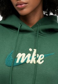 Zielona bluza z kapturem z przednią kieszenią kangurkową, z teksturowanym żółtym logo "nike" i turkusowym znakiem Swoosh. Bluza posiada sznurki do regulacji.
