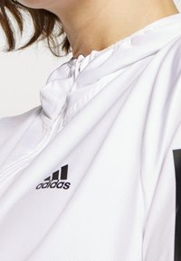 Valkoinen kevyt huppari, jossa on musta Adidas-logo vasemmassa rinnassa ja teksturoitu kangas hienovaraisella kiillolla.