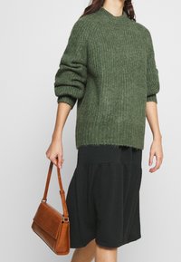 Una donna che indossa un maglione verde tessurizzato con maniche risvolte, una gonna scura a fantasia e tiene una borsa in pelle marrone per il manico.