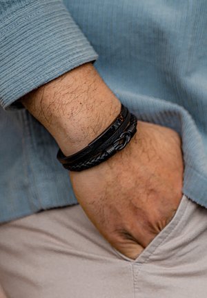 Black leather bracelet with braided texture and metal accents, worn on a wrist, contrasts with a light blue corduroy shirt.