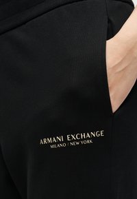Χέρι στην τσέπη μαύρου ρούχου με το κείμενο "Armani Exchange Milano / New York" τυπωμένο σε χρυσό πάνω στο ύφασμα.