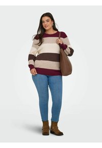 Eine Frau trägt einen gestreiften Pullover, blaue Jeans, braune Knöchelstiefel und hält eine große braune Umhängetasche. Sie steht vor einem schlichten Hintergrund.