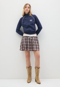 Giacca blu navy con cerniera e dettagli bianchi, gonna mini a quadri in tonalità marroni e stivali cowboy in suede beige con tacco a blocco.