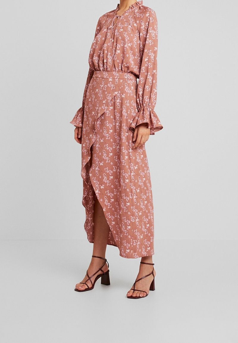 Femme portant un chemisier floral rose poussiéreux à manches longues avec des poignets à volants et une jupe midi asymétrique à volants assortie.