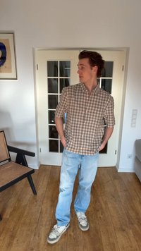 Bărbat tânăr stând în interior, purtând o cămașă maro în carouri cu mânecă scurtă, blugi albastru deschis și adidași gri, uitându-se spre dreapta cu mâinile în buzunare.