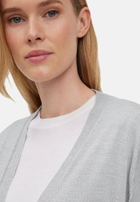 Jasnoszary kardigan z dekoltem w kształcie V, o miękkiej fakturze, noszony na białej koszulce z okrągłym dekoltem. Proste i czyste linie podkreślają warstwy.