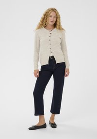 Šviesiai smėlio spalvos megztinis su apvalia iškirpte, ilgomis rankovėmis ir tamsiomis sagomis, derinamas su tamsiais trumpais džinsais ir juodais baltais bateliais.