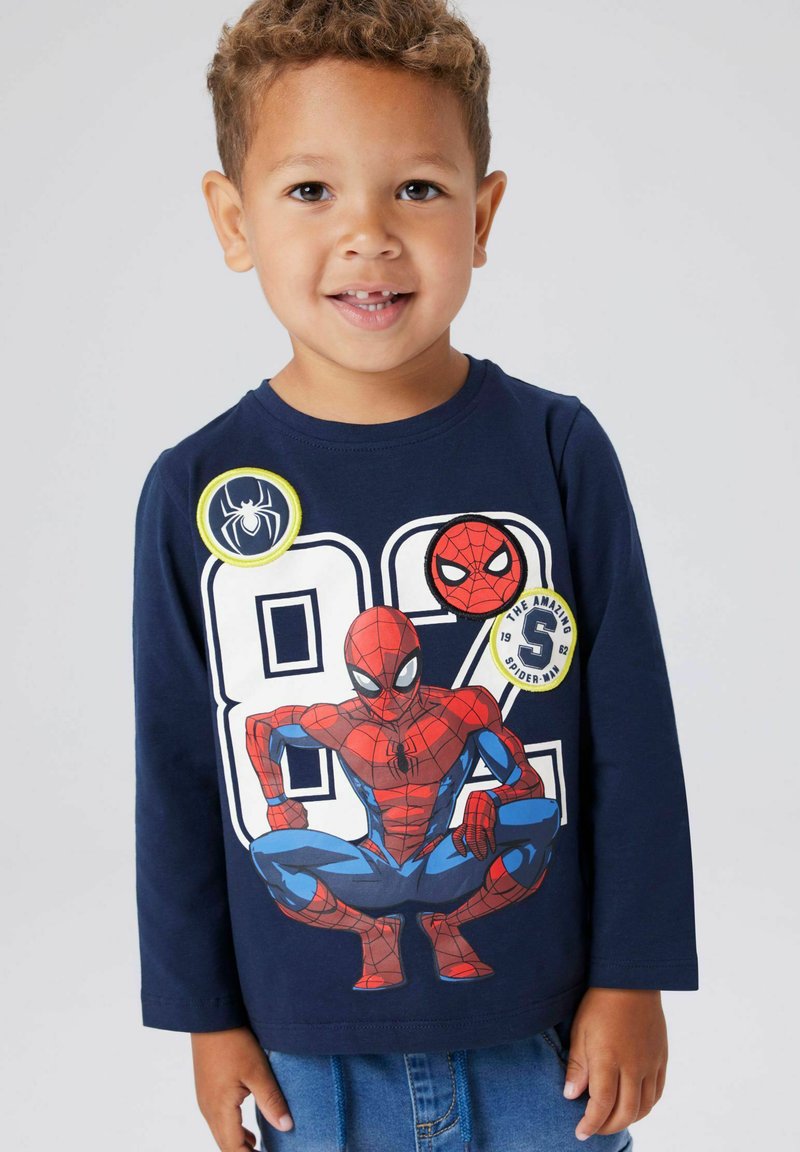 Μπλε κοντομάνικη μπλούζα με γραφικό Spider-Man, έντονα κόκκινα και μπλε χρώματα, με στρογγυλά σήματα που εμφανίζουν λογότυπα και αριθμούς Spider-Man.