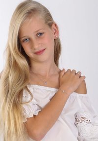 Zilveren sieraden met een hanger en een bijpassende armband, gedragen door een persoon met lang blond haar, in een witte off-shoulder top met kantaccenten.