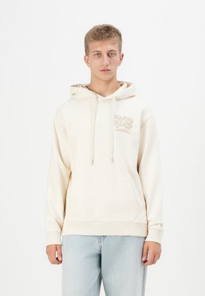 Beige hettegenser med kengurulomme, snøre og trykket logo på brystet. Laget av bomull med en myk tekstur. Kombinert med lyseblå jeans.