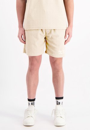 Beige Shorts mit einem strukturierten Zickzackmuster, elastischem Bund und einem Logo an der unteren Vorderseite. Getragen mit weißen Sneakern und schwarz-gestreiften Socken.