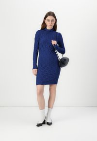 Rochie tricotată de culoare bleumarin, cu un model texturat, mâneci lungi și guler înalt. Accentuată cu o geantă neagră la umăr și șosete albe.