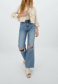 Giacca corta crema in stile uncinetto con grandi bottoni, abbinata a jeans azzurri a vita alta con strappi sulle ginocchia e sandali con piattaforma beige.