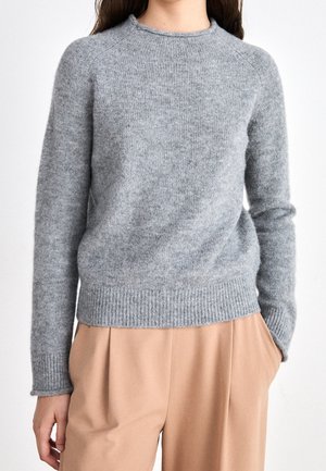 Personne portant un pull en maille gris clair avec poignets et ourlet côtelés, associé à un pantalon beige ample, une main dans la poche.