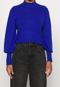 Maglione blu a maglia con collo alto e maniche a palloncino, abbinato a jeans neri a vita alta. Il materiale sembra morbido e strutturato.
