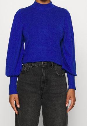 Blå strikket sweater med høj hals og ballonærmer, parret med højtaljede sorte jeans. Materialet ser blødt og tekstureret ud.