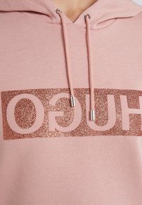 Sudadera de tela rosa claro con un gran logotipo "HUGO" en color oro rosa brillante en la parte delantera. Incluye ojales plateados y cordones a juego.