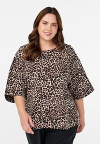 Naeratav naine lainekate pruunide juustega, kes kannab hõbedaseid rõngakõrvarõngaid ja laia leopardimustriga pluusi, mille varrukad ulatuvad küünarnukinivi, valge tausta ees.