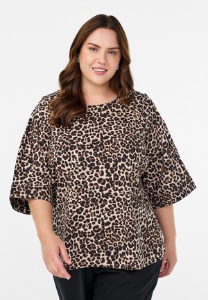 Šypsena moteris su banguotais rudais plaukais, dėvinti sidabrines žiedines auskarus ir laisvą liūto rašto marškinėlį su alkūnės ilgio rankovėmis, prieš baltą foną.