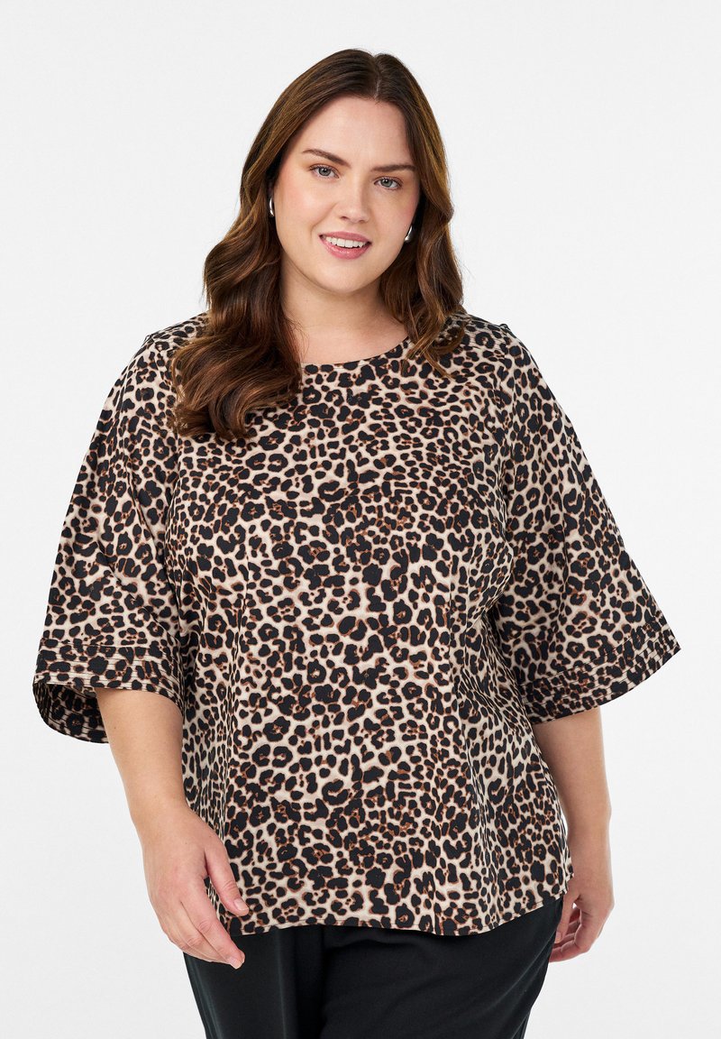 Naeratav naine lainekate pruunide juustega, kes kannab hõbedaseid rõngakõrvarõngaid ja laia leopardimustriga pluusi, mille varrukad ulatuvad küünarnukinivi, valge tausta ees.