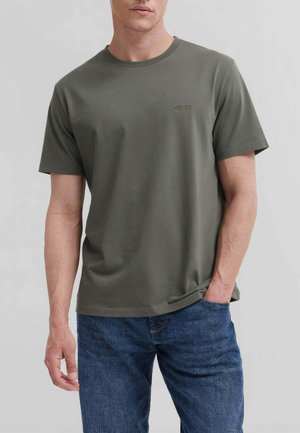 Uomo con maglietta verde oliva a maniche corte con piccolo logo sul petto e jeans blu, in piedi con una mano in tasca.