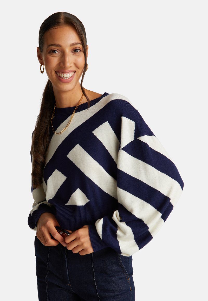 Námořnický pruhovaný oversized svetr v krémových tónech s ribbovaným lemem. Modelka má na sobě tmavé džíny, svetr má objemné rukávy a volný střih.