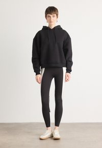 Hoodie corto nero con coulisse, abbinato a leggings neri con strisce bianche sui lati. Il modello indossa scarpe da ginnastica chiare.