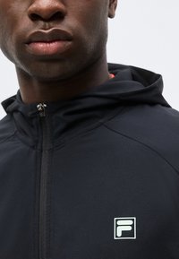 Κοντινό πλάνο ενός άνδρα που φοράει μια μαύρη φούτερ με φερμουάρ και μικρό λευκό λογότυπο «F» στο στήθος, με ουδέτερο φόντο.