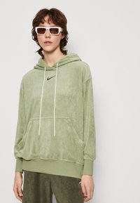 Zaļš velūra kapuce ar kenguru kabatu, auklām un izšūtu melnu Nike logo uz krūtīm. Matēta auduma ar mīkstu tekstūru.