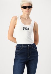 Fehér bordázott trikó, melyen a "GAP" logó található, sötétkék, magas derekú farmernadrággal párosítva, öt zsebbel és látható varrási részletekkel.