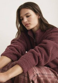 Sudadera de forro polar burdeos con cuello alto y media cremallera, que presenta un tejido texturizado y suave. Combinada con un pantalón de cuadro en tonos neutros.