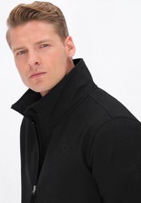 Juodas zip-up švarkas su aukštu apykakle, turintis subtilų tekstūruotą raštą ir logotipą ant krūtinės, pagamintas iš minkštos ir patvarios medžiagos.