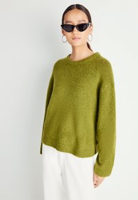 Ženska nosi oversize olivnozeleno pulover, bele hlače, črne mačje sončne očala in velike srebrne uhane v obliki obroča, stoji pred belo ozadjem.