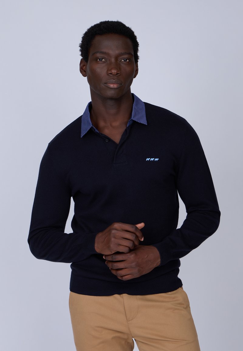Polo en maille marine avec un col à carreaux bleu texturé, arborant un petit logo animal bleu clair sur la poitrine, associé à un pantalon beige.