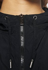 Svart lättviktsjacka med en synlig dragkedjestängning med metalliska detaljer, justerbara dragsnören vid huvan och texturerad tyg.