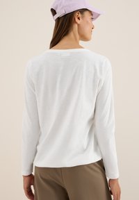 Femme portant une chemise blanche à manches longues, un pantalon marron et une casquette lilas claire, tournée le dos contre un fond beige uni.