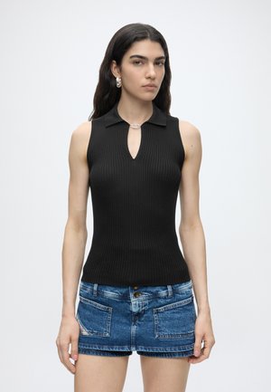 Vrouw met lang donker haar die een zwart geribd mouwloos topje met kraag en blauwe denimshorts draagt, staand tegen een effen witte achtergrond.
